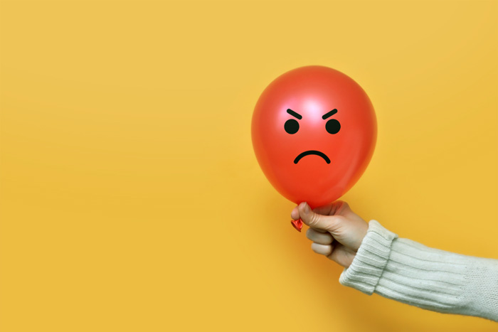 cropped shot of a person holding a red balloon that has an angry face drawn on it in black marker - all against a yellow background - anger