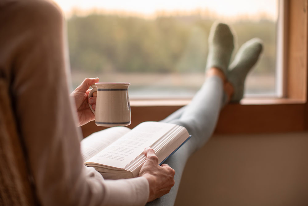 woman sitting by the window reading a book and holding a cup of coffee