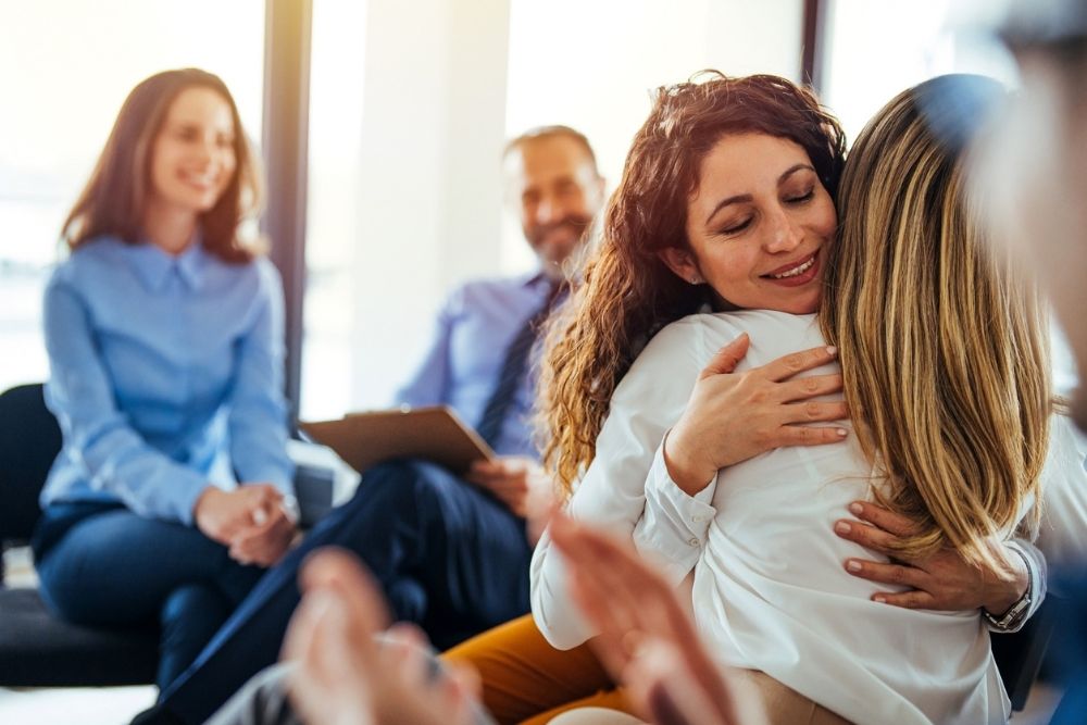 Two women hug warmly in a support group setting while others look on and smile. The atmosphere feels positive, compassionate, and encouraging.