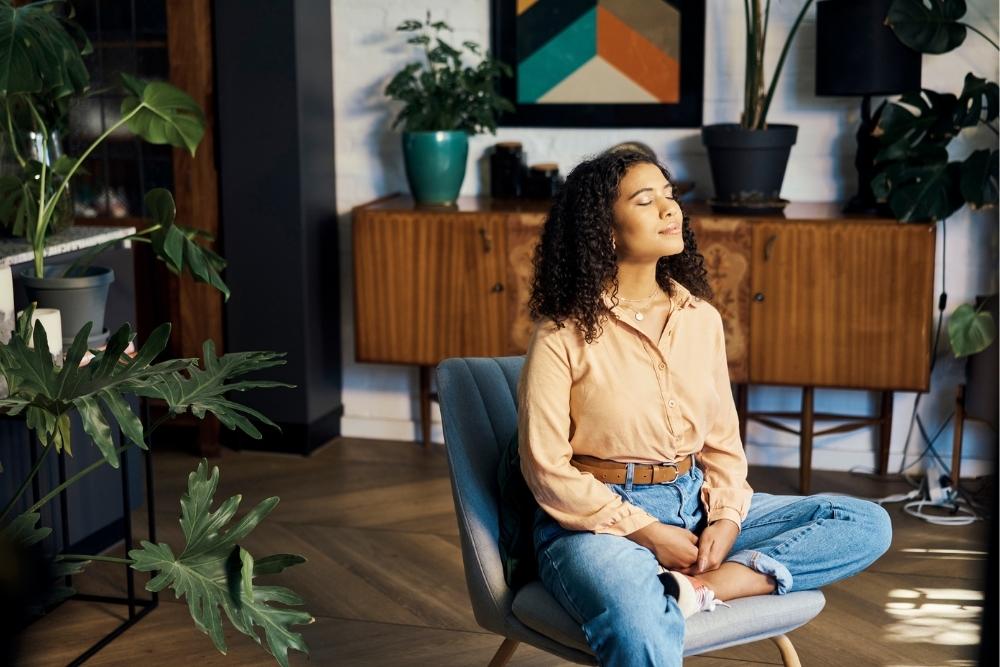 A young woman sits cross-legged on a chair in a cozy, plant-filled room, eyes closed and face relaxed as she practices mindfulness and deep breathing.