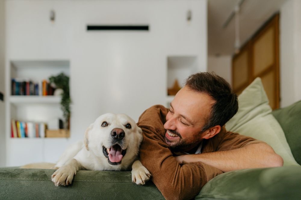 A smiling man relaxes on a couch next to his happy dog, highlighting the joy and emotional support pets can bring to everyday life.