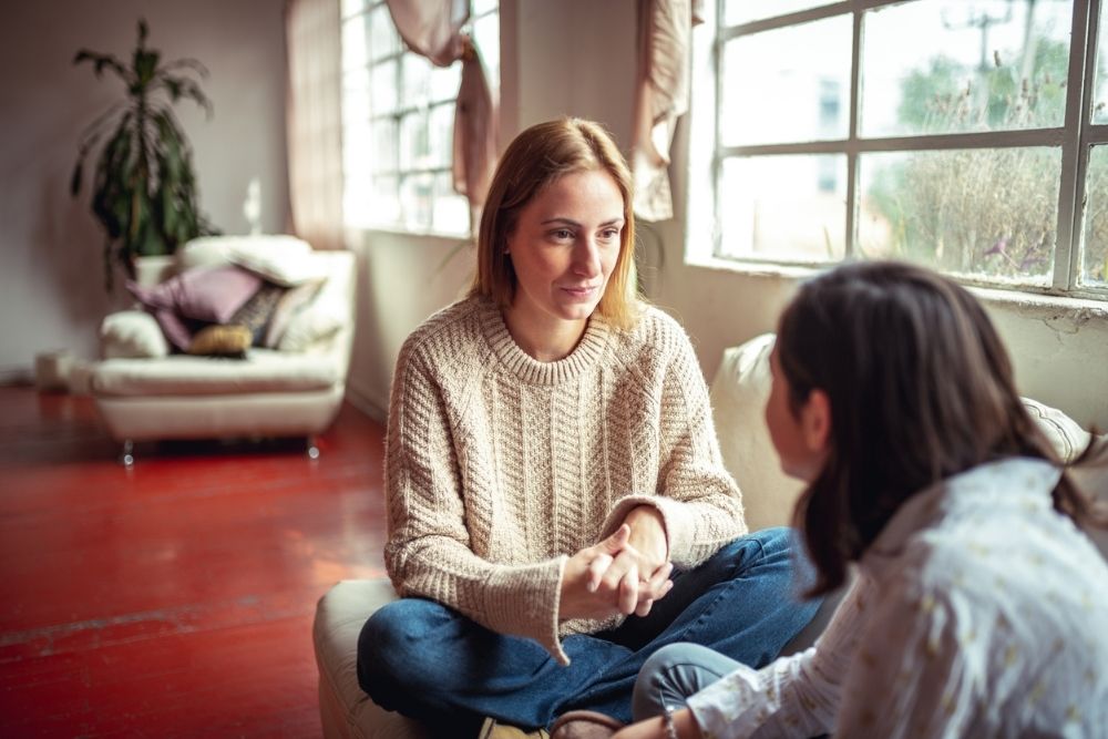 A woman sits on the floor talking to a young girl, holding her hands while having a supportive conversation in a bright living room.
