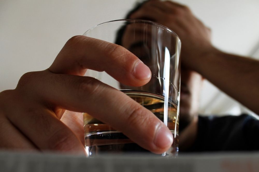 Close-up of a person holding a glass of alcohol with their other hand on their head, suggesting stress or emotional struggle.