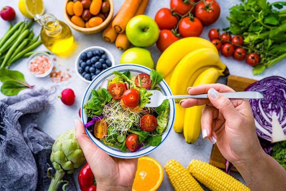 Person holding a colorful bowl of fresh salad surrounded by fruits and vegetables, symbolizing healthy eating and nutritional recovery after substance use.
