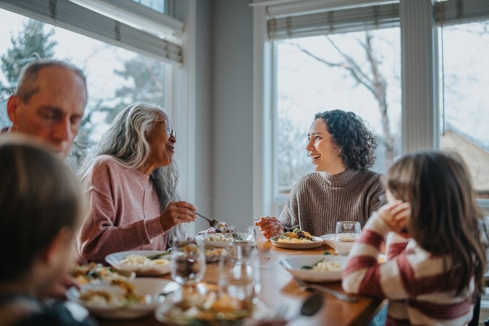 A multigenerational family shares a warm meal together at a dining table, smiling and talking in a bright room with large windows.