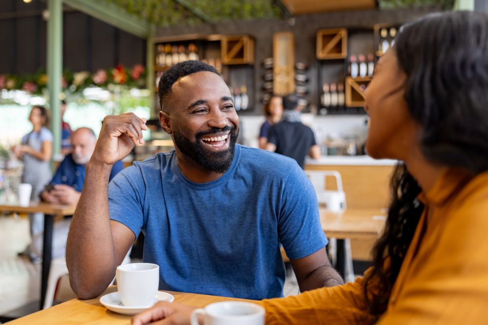 A man and woman sit together at a café, smiling and laughing while enjoying coffee, symbolizing joy, connection, and emotional well-being.