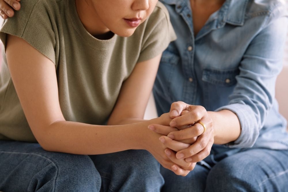 A person sits with their hands clasped, looking down, while another person beside them gently offers support with a hand on their shoulder. The scene conveys comfort, empathy, and emotional support.