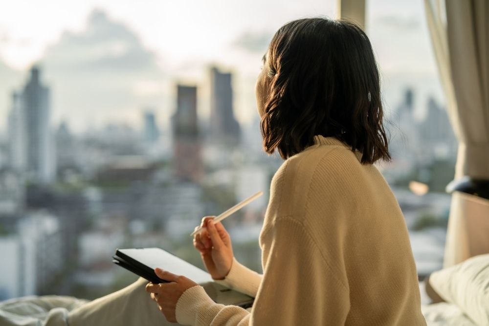 A woman sits by a window overlooking a city, holding a notebook and pen while reflecting quietly.