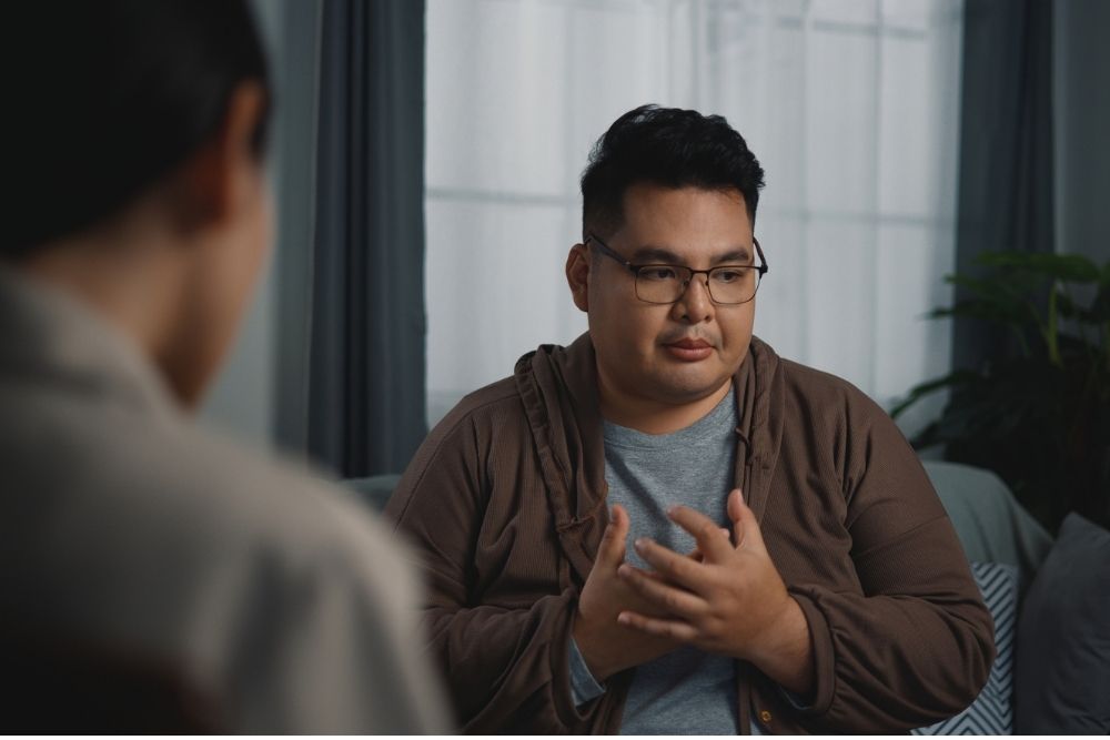 A man sits in a therapy session, speaking with a counselor and gesturing with his hands in a calm, supportive outpatient setting.