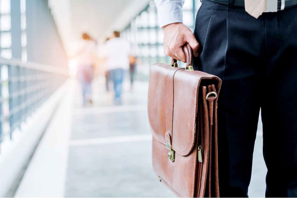 A professional holding a leather briefcase while walking through a bright, modern hallway, with people blurred in the background.