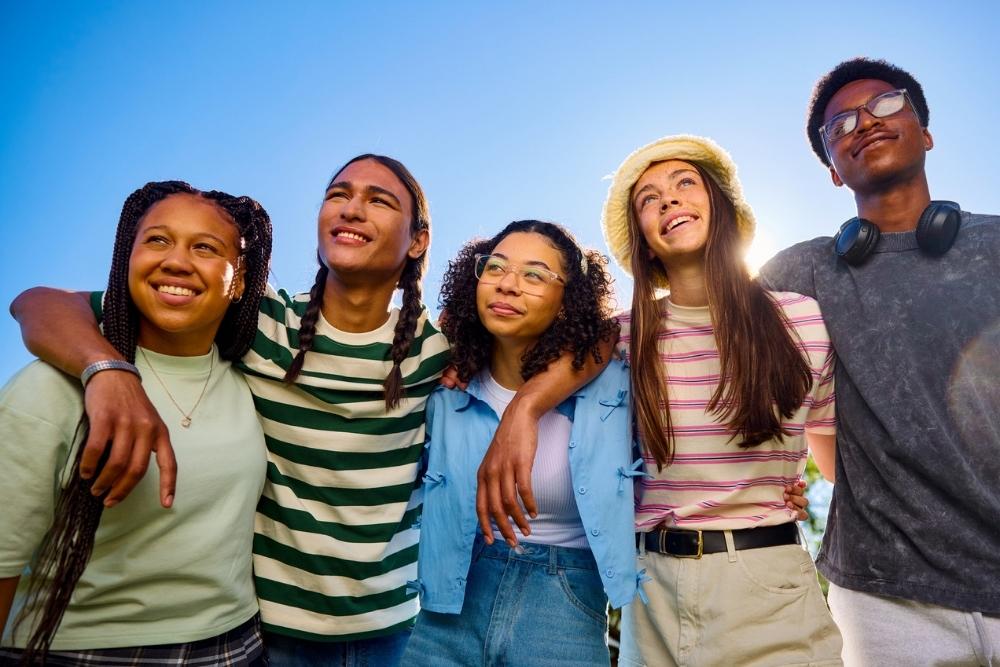 A diverse group of five smiling friends staying healthy in recovery under a clear blue sky. They appear joyful and relaxed, exuding a sense of warmth and togetherness.