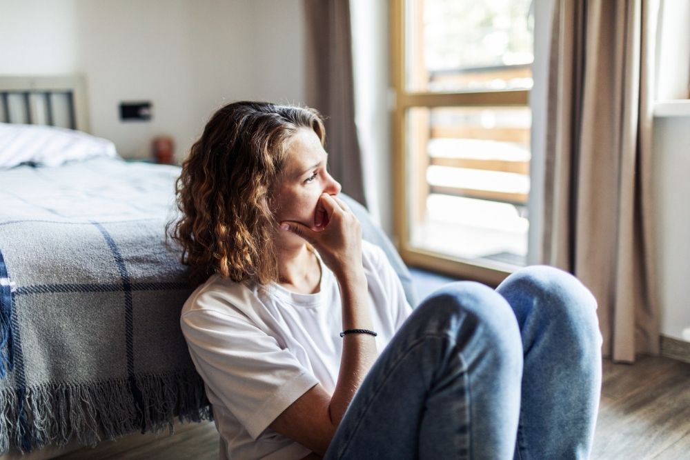 A person sits on the floor by a bed, looking thoughtfully out a window with their hand resting on their chin, lost in the depths of complicated grief.